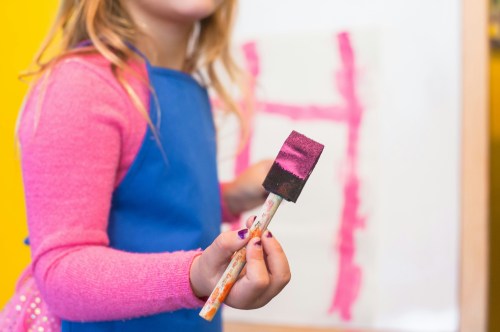 Closeup of a young girl wearing a blue apron and holding a foam paintbrush dipped in hot pink paint.
