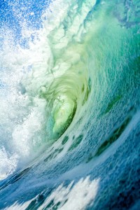 A close-up view of a curling ocean wave, showcasing vibrant shades of green and blue water and frothy white spray.